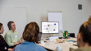 Gruppe sitzt am Tisch und sieht eine Präsentation auf einem Bildschirm