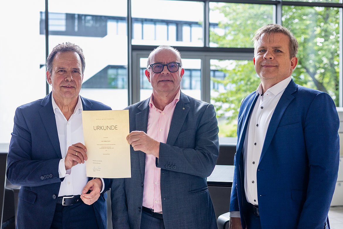 Rektor und zwei Professoren stehen nebeneinander mit einer Urkunde in der Hand.