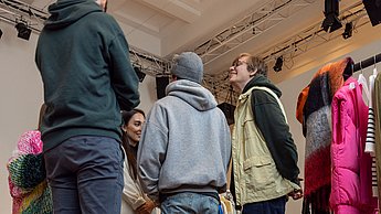 Kleidung liegt zusammengestellt auf dem Boden und Studierende stehen abgewandt dahinter
