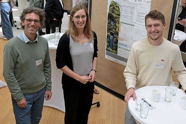 Drei Personen an Stehtisch vor Poster