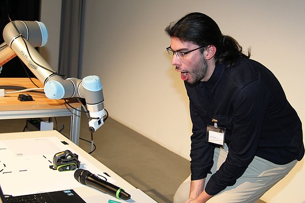 Mitarbeiter steht vor Roboterarm und demonstriert den Zuschauern etwas.