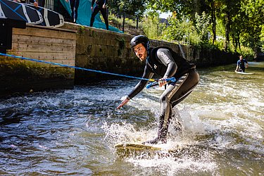 Zu sehen ist eine Person im Neoprenanzug. Sie steht auf einen Surfbrett auf der Enz und hält sich an einem Seil fest.