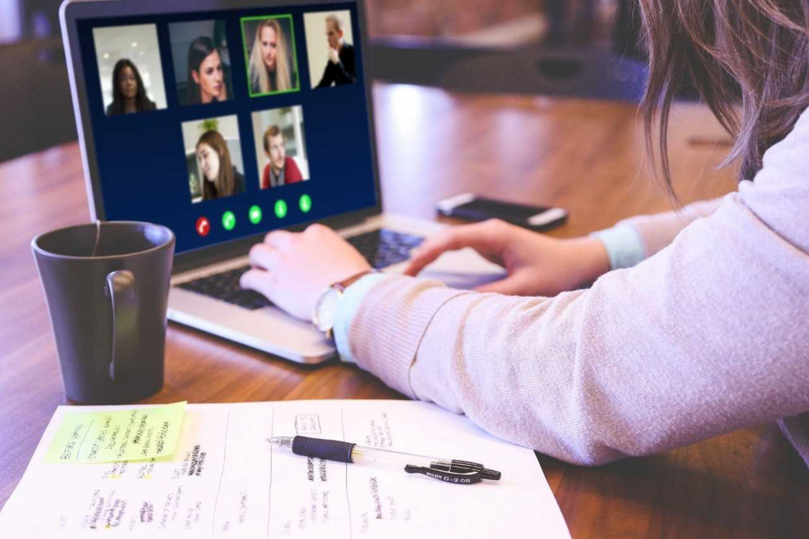 Das Bild zeigt einen Laptop mit einer geöffneten Videokonferenz und einen Teil des Oberkörper einer Frau mit braunen Haaren. Vor dem Laptop befinden sich Unterlagen, ein Stift und eine Tasse. 