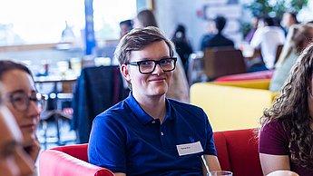Ein Student lächelt in die Kamera und sitzt auf einem Sofa.