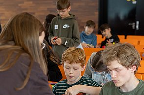 Zwei Jungen basteln an Gesichtern mit Schnippseln und werden dabei betreut von einer studentischen Hilfskraft