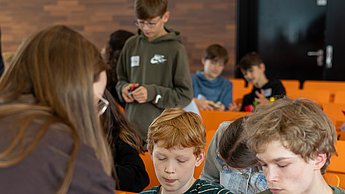 Zwei Jungen basteln an Gesichtern mit Schnippseln und werden dabei betreut von einer studentischen Hilfskraft