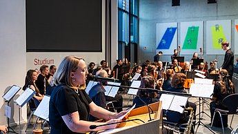Frau am Rednerpult beim Sprechen, im Hintergrund ein Orchester (Menschen mit ihren Instrumenten)