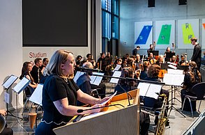Frau am Rednerpult beim Sprechen, im Hintergrund ein Orchester (Menschen mit ihren Instrumenten)