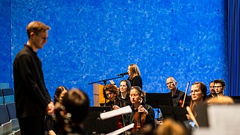 Das Orchester der Hochschule Pforzheim in Aktion, im Hintergrund ist eine Frau (die Sprecherin) am Mikro zu sehen
