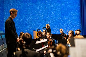Das Orchester der Hochschule Pforzheim in Aktion, im Hintergrund ist eine Frau (die Sprecherin) am Mikro zu sehen
