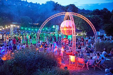 Many people can be seen in a park. In the center is an open dome with a disco ball. Everything lights up colorfully.