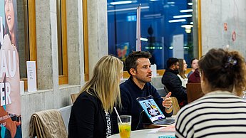 Eine Frau und ein Mann an einem Tisch mit Studierenden