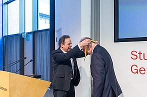 Der Rektor hängt dem Professor eine Medaille um den Hals.