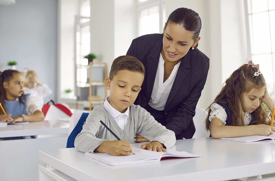 Kinder in Schulklasse; eine Lehrerin beugt sich zu einem Jungen, der schreibt.