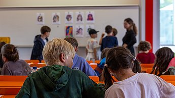 Zwei Jungen basteln an Gesichtern mit Schnippseln, im Hintergrund sieht man Beispielfotos, die die Aufgabe verdeutlichen sollen