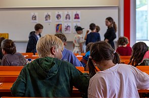 Zwei Jungen basteln an Gesichtern mit Schnippseln, im Hintergrund sieht man Beispielfotos, die die Aufgabe verdeutlichen sollen