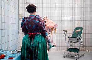 Zwei kostümierte Personen stehen in einem weißen Raum. Im Hintergrund steht ein Stuhl während vorne ein Tisch mit Operationswerkzeug steht