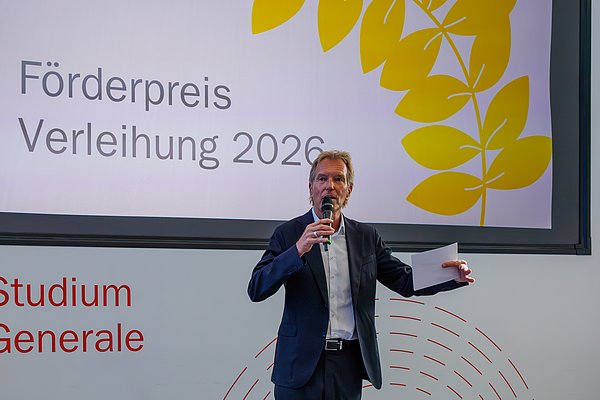 Mann mit Mikro steht vor Präsentation mit Schriftzug Förderpreisverleihung 2026