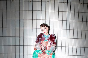 Person in buntem Kostüm steht in einem sauberen, weißen Zimmer vor einem Tisch