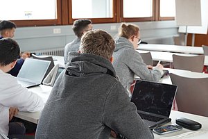 Ein Studierender sitzt mit dem Rücken zur Kamera in einem Hörsaal mit einem Laptop vor sich