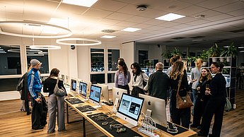Menschengruppe steht vor einem Tisch auf dem viele Computer stehen. Auf den Bildschirmen werden Bilder gezeigt.