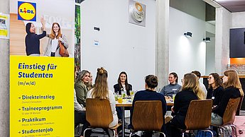 Ein Roll-Up der Firma Lidl und Personen an einem Tisch im Gespräch