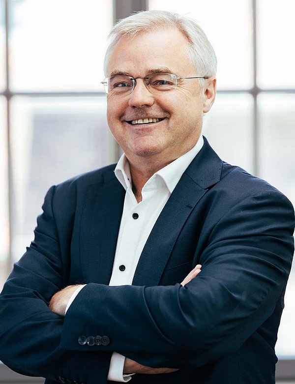 Älterer Herr mit weißem Haar (Ralf Otte) lächelt mit verschränkten Armen in die Kamera.