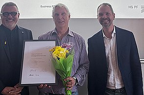 Prof. Dr. Thomas Cleff mit Preisträger Prof. Dr. Kevin Johnston, der eine Urkunde und Blumen in der Hand hält, sowie Prof. Dr. Florian Haas