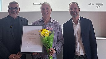 Prof. Dr. Thomas Cleff mit Preisträger Prof. Dr. Kevin Johnston, der eine Urkunde und Blumen in der Hand hält, sowie Prof. Dr. Florian Haas