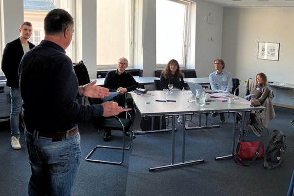 Foto vom Workshop, wo eine Person referiert und mehrere Personen um einen Tisch herum sitzen