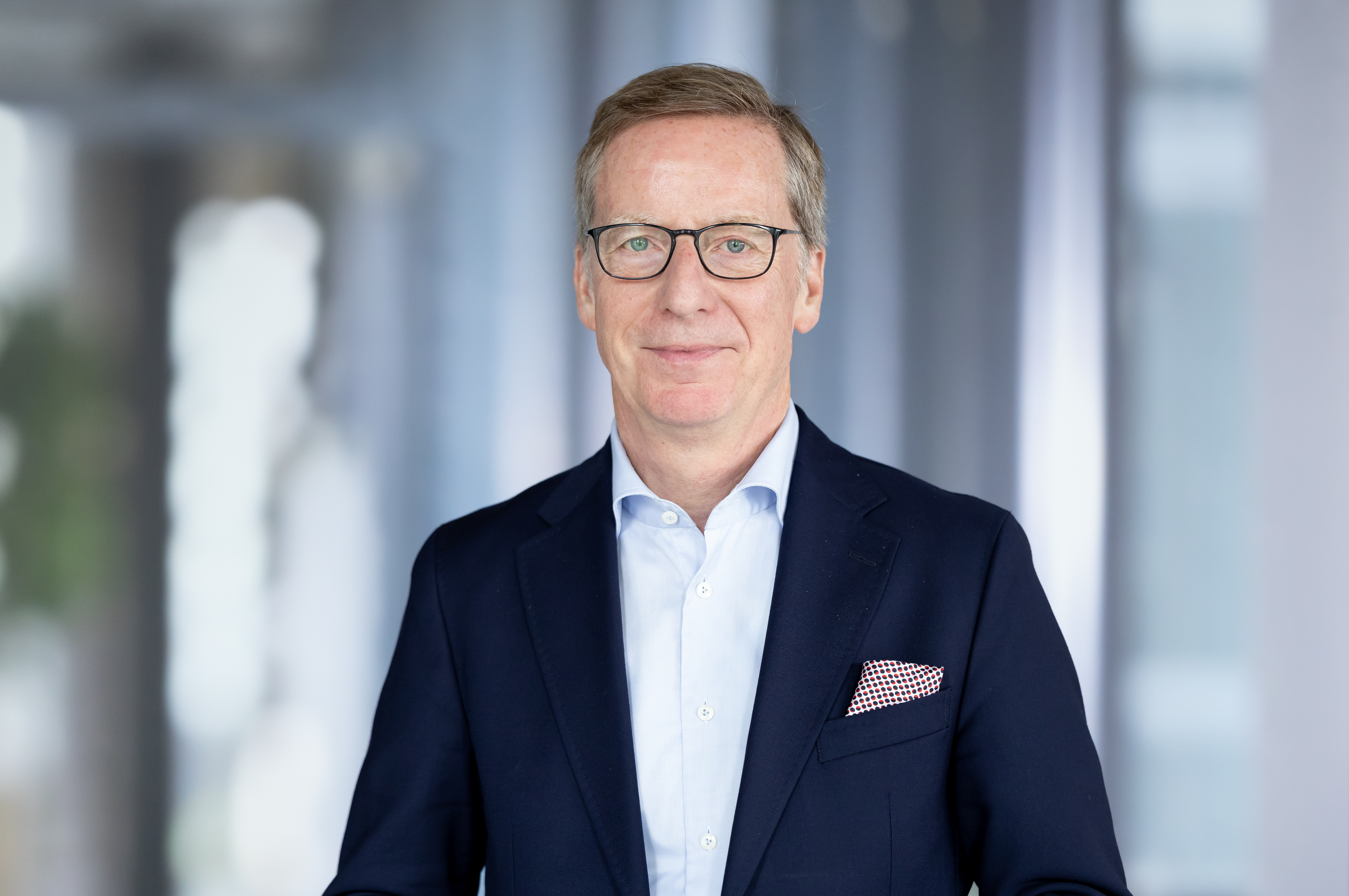 Mann im Anzug mit grauem Haar und Brille schaut in die Kamera.